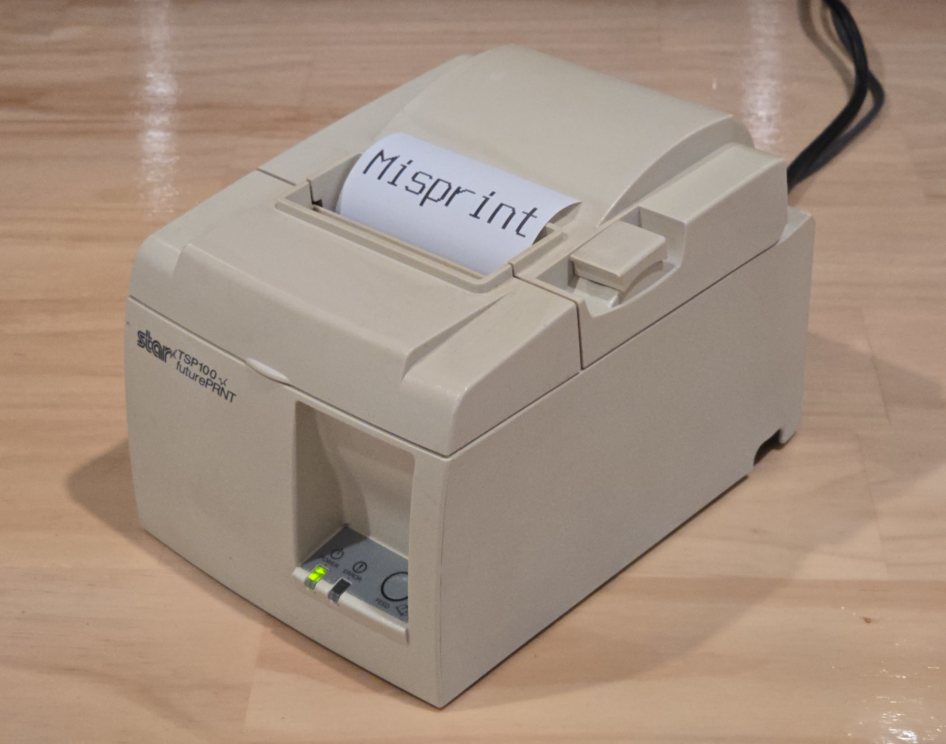 A photo of the Star Micronics TSP100 printer with a small amount of receipt paper spooled out the top that has the text "Misprint" printed on it.